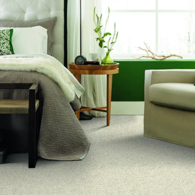 Bed room with grey carpet, plan on wooden night stand, and a green window side. theres beige accents all around, with a cream corner chair, and grey bedding.